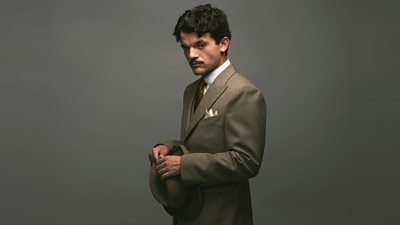 First look photo of Edward Bluemel as Hercule Poirot. Dressed in a suit and tie, he holds a bowler hat in both hands and looks to the camera. 
