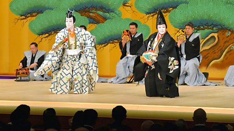 Two characters stand on stage, dancing whilst wearing elaborate costumes and make-up indicative of an Asian location.