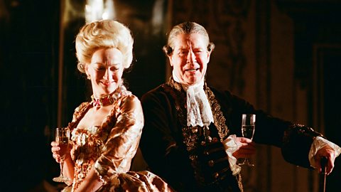 A man and woman stand back to back, laughing whilst holding a drink each. They are wearing elaborate period costumes and wigs.