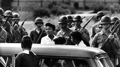 A black and white photograph of some of the Little Rock Nine students arriving at school, surrounded by soldiers holding weapons.