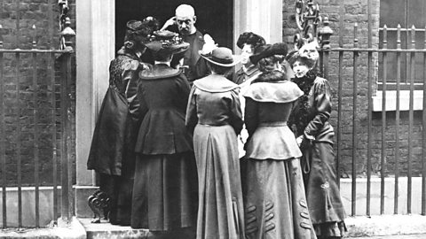 A group of suffragettes gather outside a house. 