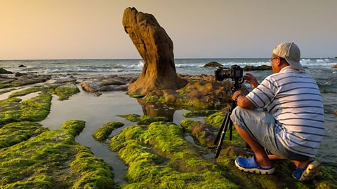 A camera operator filming on the shore at dusk.
