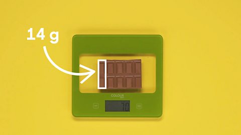 A chocolate bar on a set of scales, where a chunk of it is labelled as weighing fourteen grams