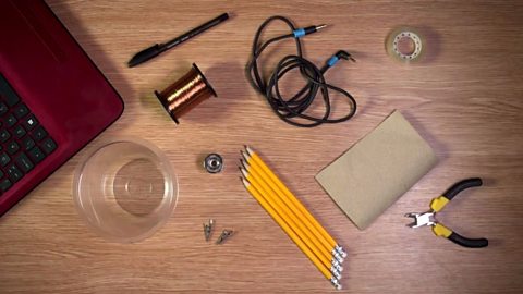 An aerial shot of a pen, pencils, crocodile clips, insulated wire, wire cutters, sandpaper, speaker jack cable and a plastic cup