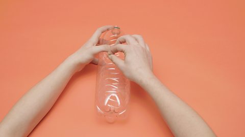 A large plastic bottle the person is using a pin to make 3 small holes at different heights of the bottle its advised to use the pencil to widen the holes.