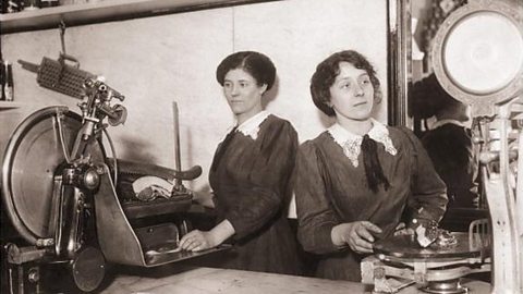 Female shop assistants slice meat and weigh it for a customer in the early 1900s