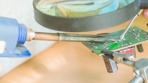 Someone holding a magnifying glass and a soft soldering pen working on a circuit board.