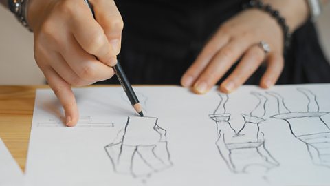 A pair of hands shown drawing a stretch of figures with a black pencil on white paper.
