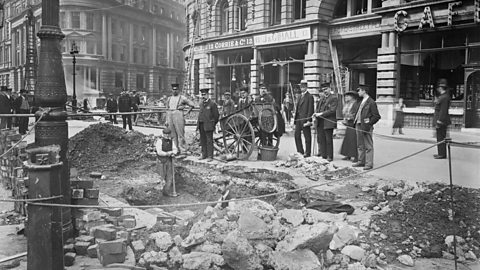 A group of men stand around a bomb damaged street the day after an air raid during World War One