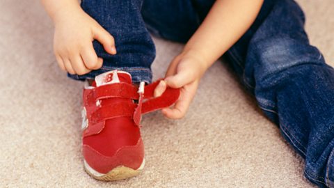 A young child sat on the floor fastening their red Velcro trainers.