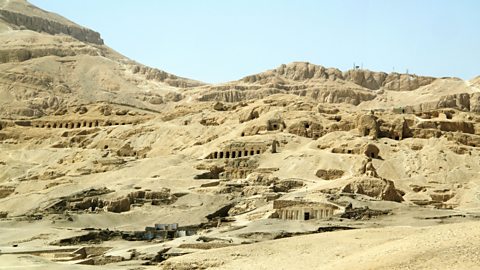 A photo of a wide valley with lots of tombs built into the sandy edges. Lots of small dark entrances are visible in the picture.