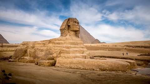 A photo of a limestone sphinx in front of the pyramids of Giza. The sphinx has huge front paws and it's head is carved to look like a man wearing a royal head cloth.