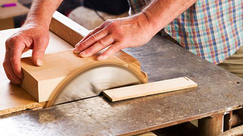 A man’s hands carefully push a piece of timber against a circular saw to cut it.