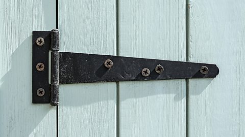 A large, black, metal T-hinge holds a light blue painted wooden gate in place.