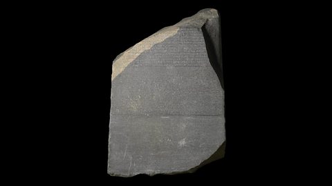 A photo of the famous Rosetta Stone. It is a large, dark grey piece of granite with rough edges. It has inscriptions carved into it.