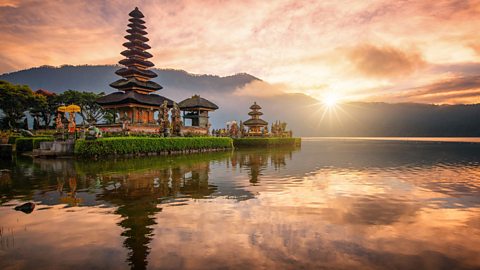 A traditional religious temple against a low sun setting in the background, casting light over the surrounding water.