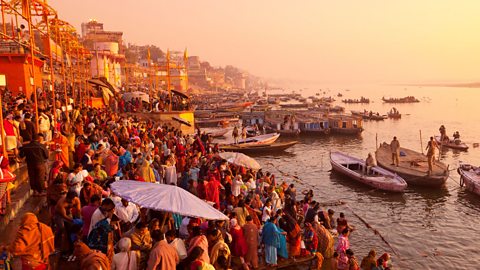 A crowd gathered at the edge of a river. Multiple boats are docked nearby and large umbrellas provide shade to some groups and individuals. 