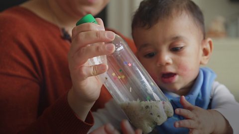 Baby boy looking at a shaker made of a rice-filled bottle.