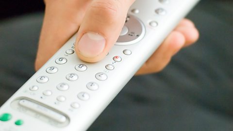 A hand holds a remote control for a television, pressing the zero button.