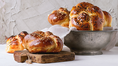 Multiple pieces of bread, placed on a wooden plank and in a metal bowl, baked into circular knotted shapes. 