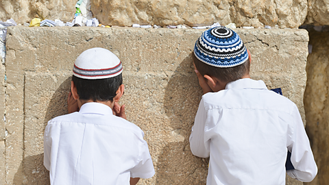 Two young boys, wearing brimless caps, cover their eyes and rest their head against the same stone wall.