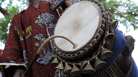 A talking drum player playing the drum in traditional dress. 