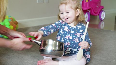 A little girl saying 'bang' as she is handed a saucepan.