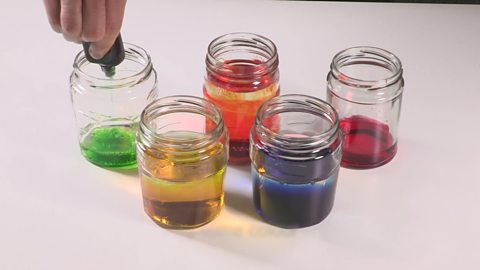 5 jam jars filled with different coloured water, the last one is having food colouring added to it.