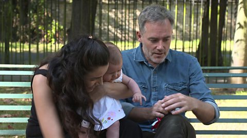 A mum and dad sat on a bench with their daughter.