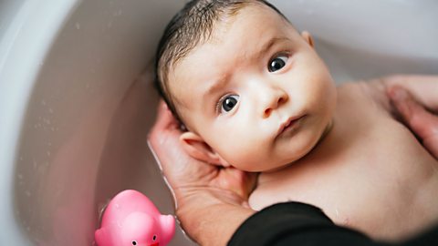 Baby in a bath with a pink rubber duck being held as it is bathed 