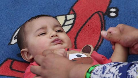 A baby boy screwing up his face and pulling away as his mum holds his hands.