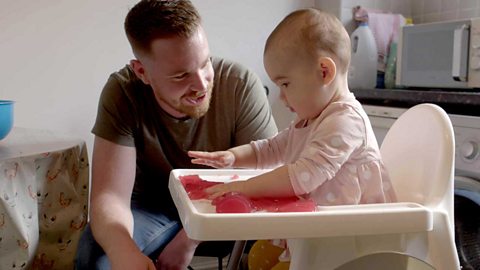 A dad with his daughter at a high chair playing with a pile of red jelly.