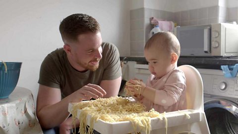 A dad with his daughter at a high chair playing with a pile of cooked spaghetti.