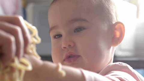 A baby girl looking at some cooked spaghetti in her hand.