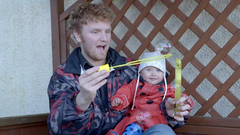 A little girl sitting on her big brother's lap as he blows bubbles.