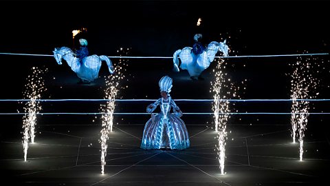 A woman in an elaborate and illuminated period dress stands on stage beneath several exploding firework pyrotechnics.