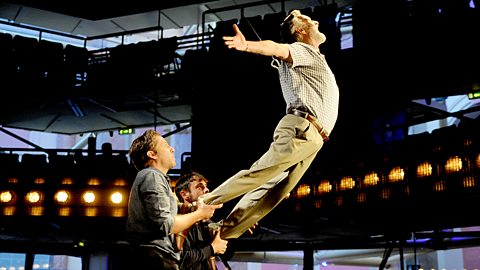 A man is supported by two strings from the top and two men from beneath as he practices 'flying' on stage.