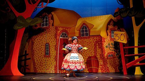 A dame is on stage in a pantomime performance - they are wearing elaborate and colourful costume and stand amongst a bright village set.