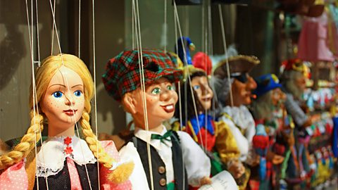 A row of marionette puppet characters, with one male and one female character in focus at the front, hang by their strings backstage.