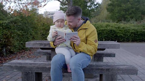 The same dad and his toddler daughter looking at a book on a park bench.