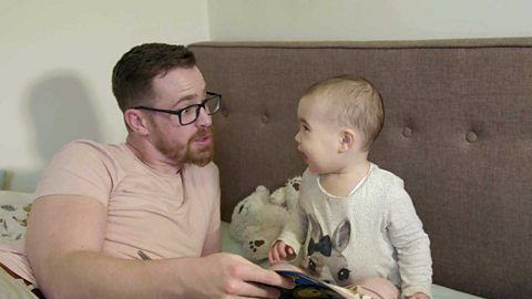 A dad and his toddler girl looking at a book on a bed.