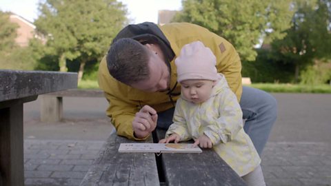 A toddler girl pointing to a picture in a book as her dad looks on.