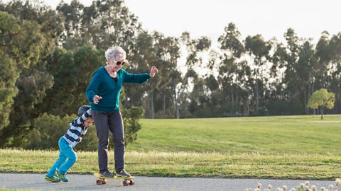 A grandmother plays on a skateboard with her grandchild who is helping her