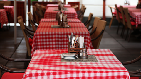 A photo of a table in a restaurant. The table has a red and white table table cloth, four plates, salt, pepper and cutlery.