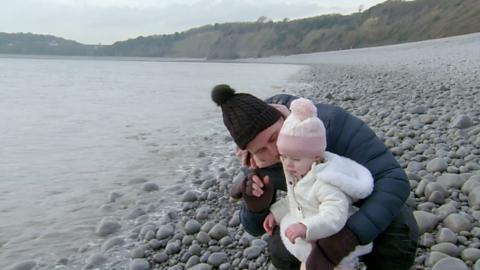 On a pebble beach a man holding his daughter and pointing with his hand