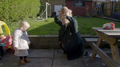 In a garden, woman holding a ball on her head while her daughter looks on