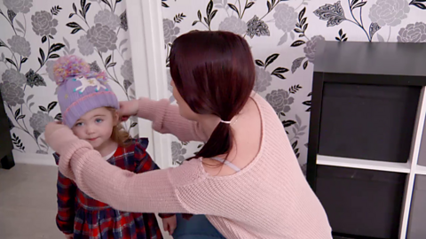 Woman in pink jumper putting purple bobble hat on toddler