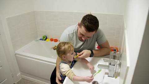 Dad helping his toddler brush her teeth at bathroom basin