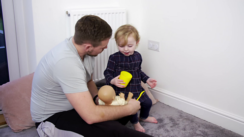 Dad holding doll while his toddler holds a cup to its mouth