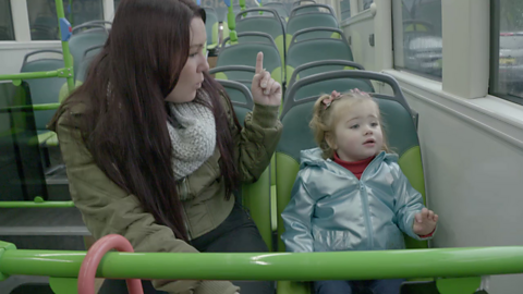 Woman and toddler sat next to each other on bus, woman is pointing